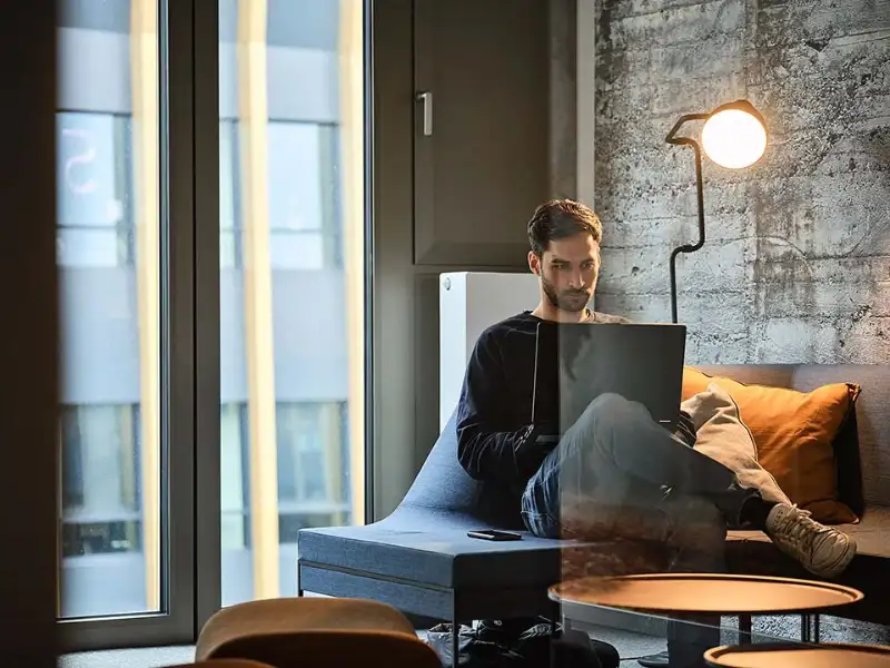 Ein Mann sitzt mit überschlagenen Beinen auf einer Couch im isolierten Büro und arbeitet am Laptop Ein Mann sitzt mit überschlagenen Beinen auf einer Couch im isolierten Büro und arbeitet am Laptop